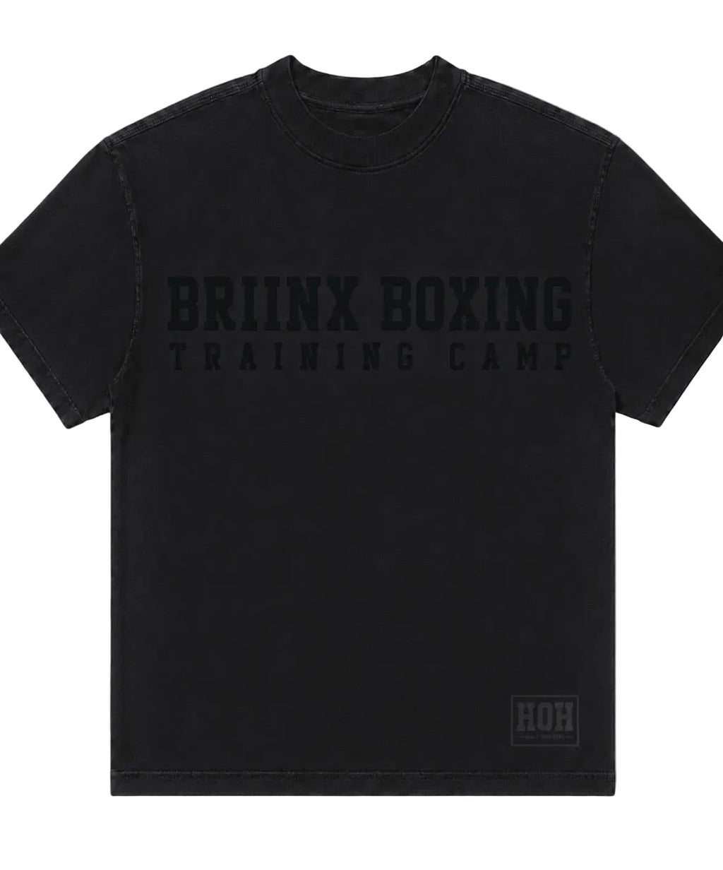 A black short-sleeve t-shirt with 'BRIINX BOXING TRAINING CAMP' printed in a tonal black font across the chest, and a small 'HOH' logo patch on the bottom right hem, shown against a neutral gray background.