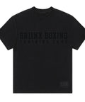 A black short-sleeve t-shirt with 'BRIINX BOXING TRAINING CAMP' printed in a tonal black font across the chest, and a small 'HOH' logo patch on the bottom right hem, shown against a neutral gray background.