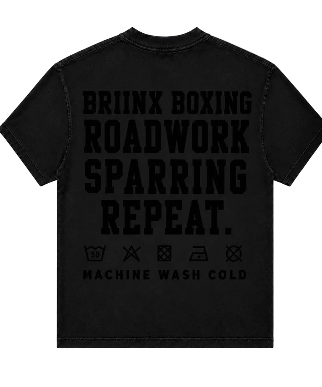 A black short-sleeve t-shirt shown from the back, featuring the words 'BRIINX BOXING ROADWORK SPARRING REPEAT.' in large black block letters, with care symbols and 'MACHINE WASH COLD' printed below.